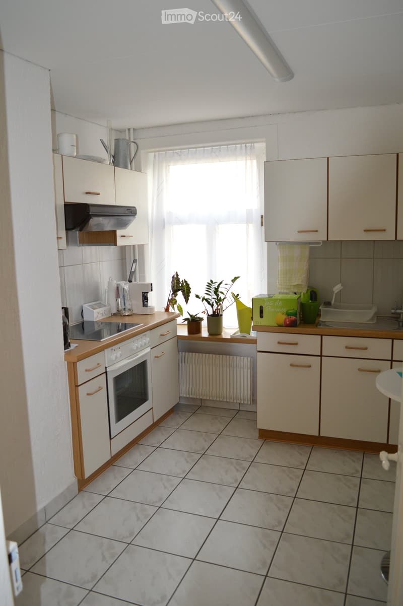 A small, neat kitchen with white tiles, white cabinets, built-in appliances, and a wooden countertop. There is a white porcelain sink with a faucet, a window with white curtains, and a potted plant on the window sill. A white towel is hanging on the oven.