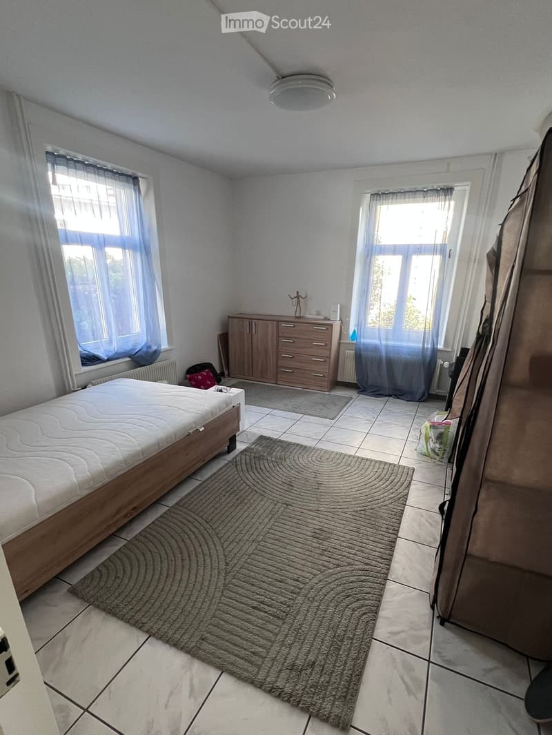 Bright bedroom with a wooden bed, a wardrobe, a large rug, and two windows with curtains. The room features tiled flooring and a radiator under each window.