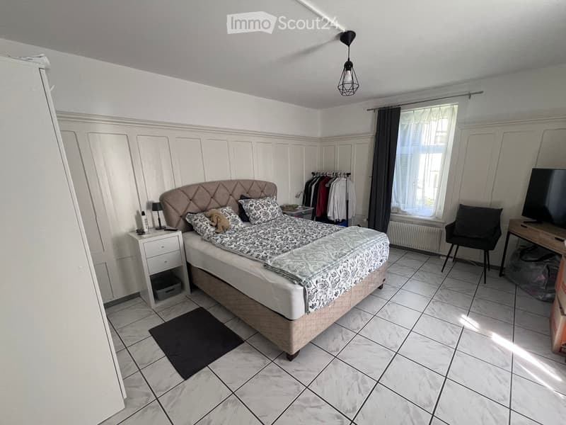 Cozy bedroom with a large bed, nightstand, desk, chair, and mirror. The room features tiled flooring and large windows with curtains.