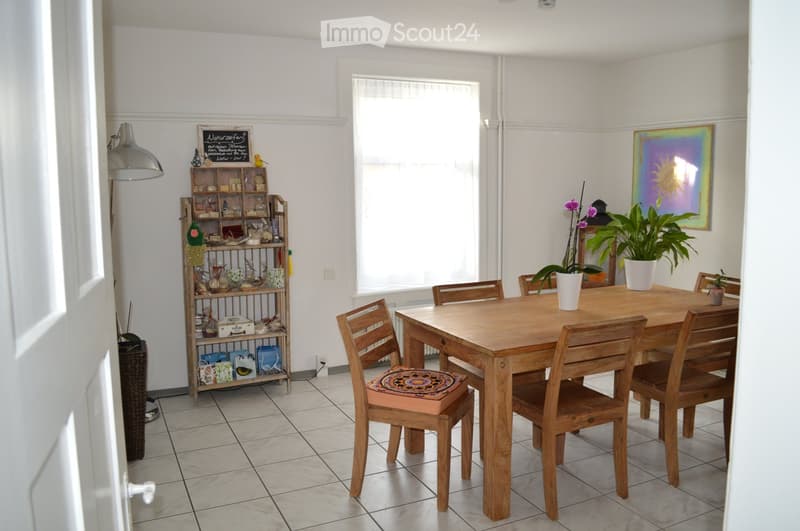 A bright dining area with wooden furniture, white walls, and white tile flooring. The dining area features a wooden table and wooden chairs with cushioned seats. There is a shelf with various items on the left side and a painting and plants on the right s