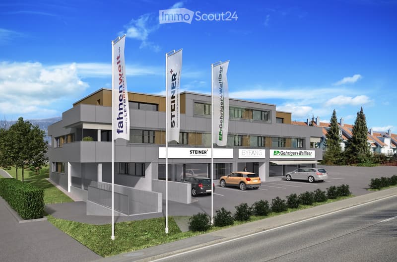 Modern two-story building with a gray facade, large windows, and a parking area with several cars. The building has multiple company logos displayed on the facade and flags.