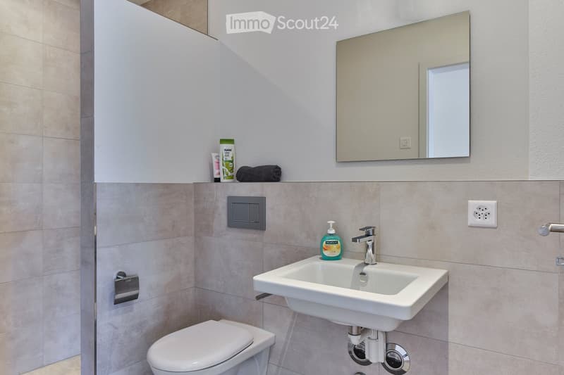 Modern bathroom with a white sink, chrome faucet, toilet, and a large mirror above the sink. Beige tiles and a towel on the shelf.