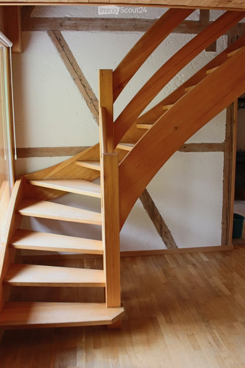 Corridor / Hallway. Wooden staircase with open wooden railings and steps, leading up to an upper level. The walls are white plaster with visible wooden beams, and the floor is made of light wooden planks.