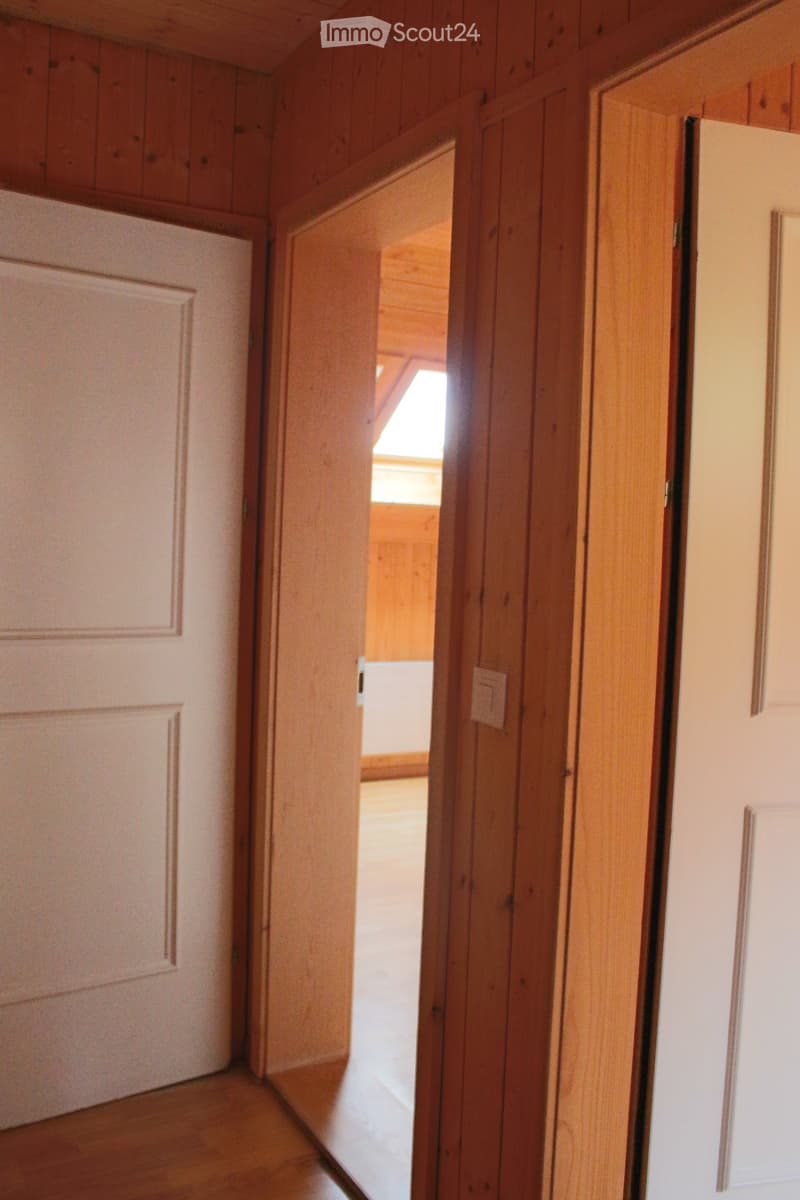Corridor / Hallway. A narrow wooden hallway with white walls, featuring a partially open door leading to another room with wooden walls and a slanted ceiling.