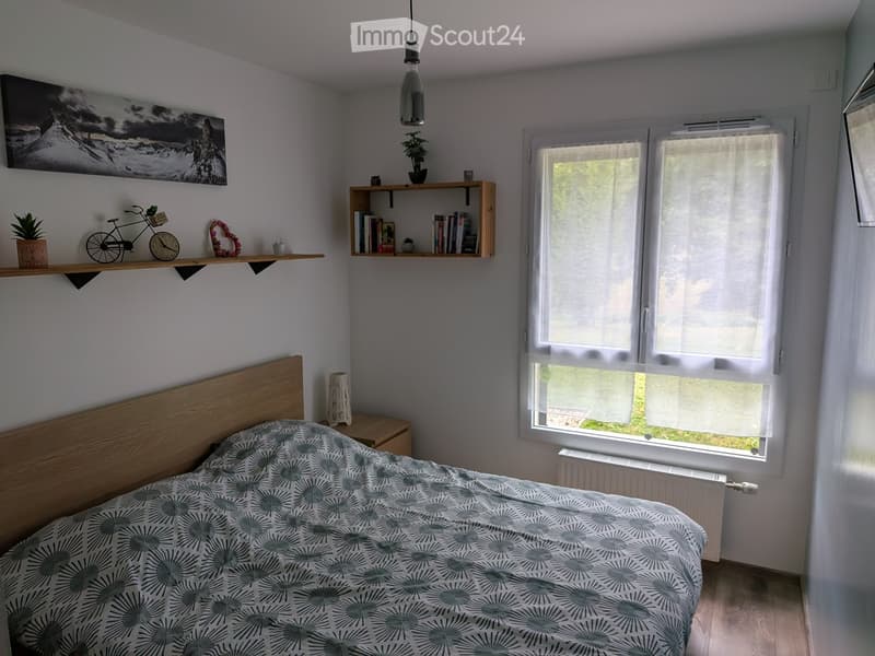 Cozy bedroom with a wooden bed, patterned bedspread, nightstand, shelves with books and decorative items, and large windows with blinds.