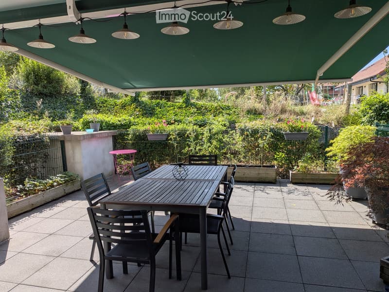 A covered outdoor patio with a large wooden dining table, six chairs, and a small pink stool, surrounded by greenery and potted plants.
