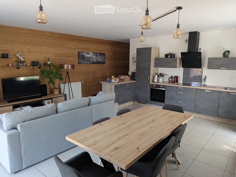 Open-plan living and dining area with a grey sofa, wooden dining table, chairs, modern kitchen with grey cabinets, oven, and pendant lights.