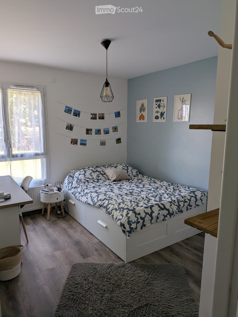 Cozy bedroom with a single bed, patterned bedding, a small table with a chair, a hanging pendant light, and several picture frames on the wall.