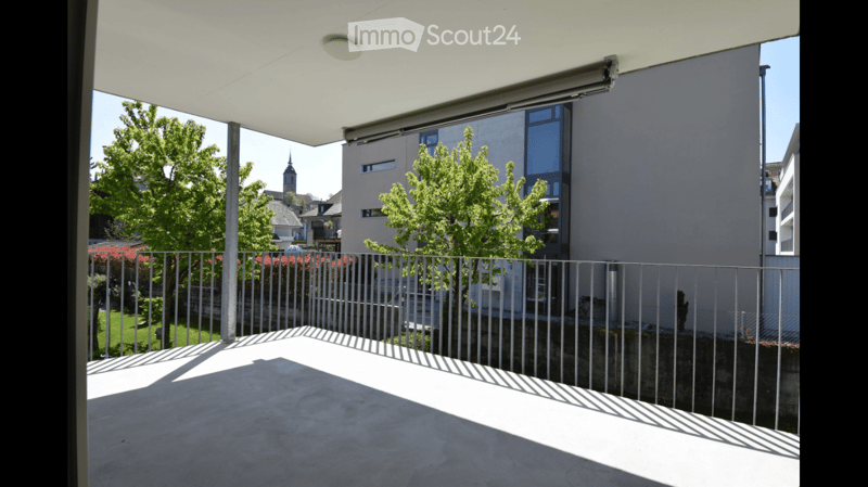 Modern balcony with a white roof, metal railing, and a view of a cityscape with trees and buildings.