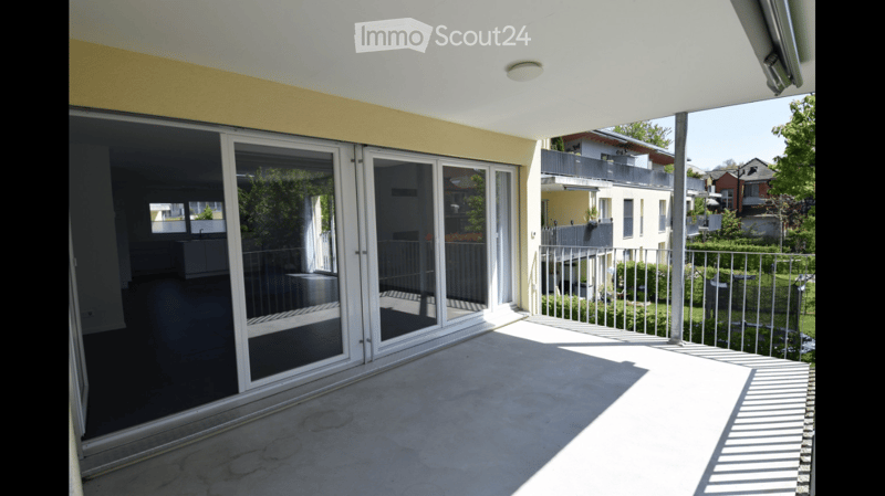 Spacious balcony with large glass sliding doors, concrete floor, metal railing, and view of neighboring buildings and greenery.