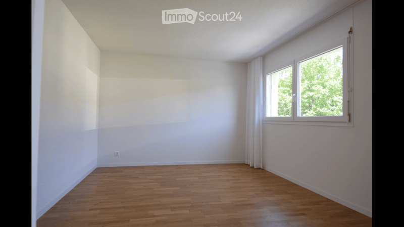 Empty room with wooden flooring, white walls, and a large window with white curtains.