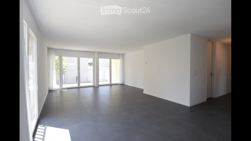 Empty room with white walls, dark tiled floor, large windows, and sliding glass doors leading to a balcony.