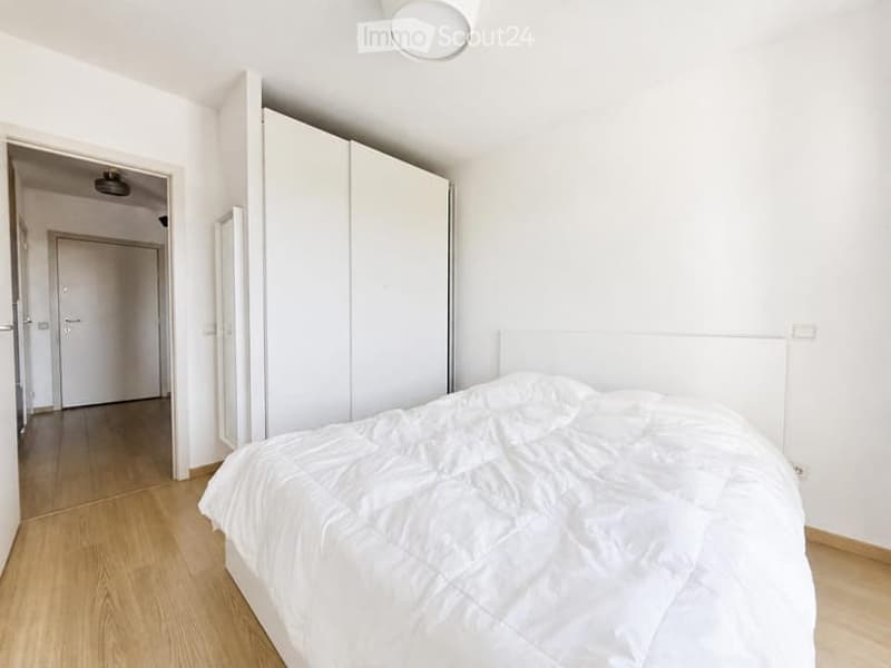Bright, minimalist bedroom with white walls, wooden flooring, a neatly made bed, and a large white wardrobe.