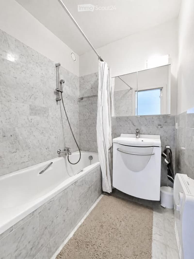 Modern bathroom with a white bathtub, shower curtain, sink, mirror, and cabinet. The walls are tiled in a light grey marble pattern, and a small towel is visible on the floor.