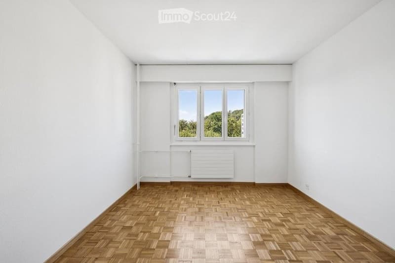Completely empty room with white walls, wooden herringbone flooring, and large windows with a view of greenery.