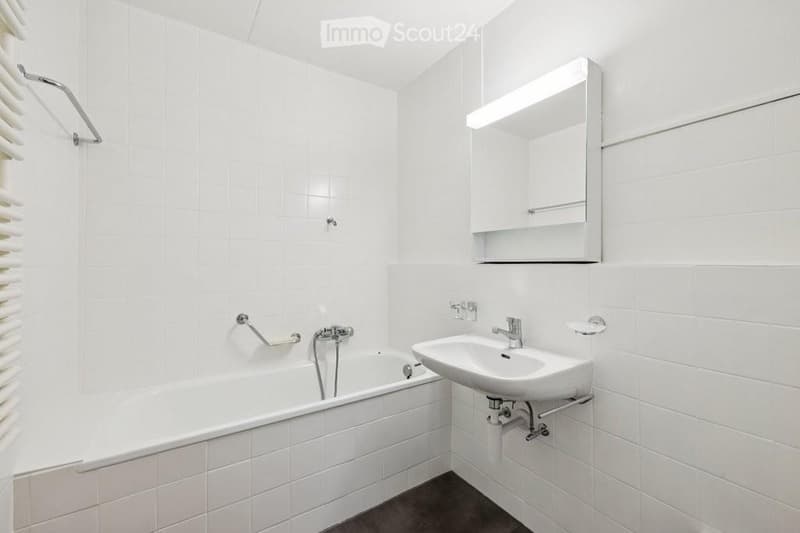 Modern bathroom with white tiled walls and floor, featuring a bathtub, sink, mirror, and built-in storage cabinet.