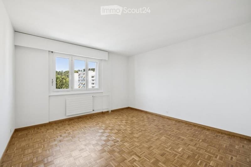 Completely empty room with white walls, wooden parquet flooring, and large windows with white frames. No furniture or equipment present.