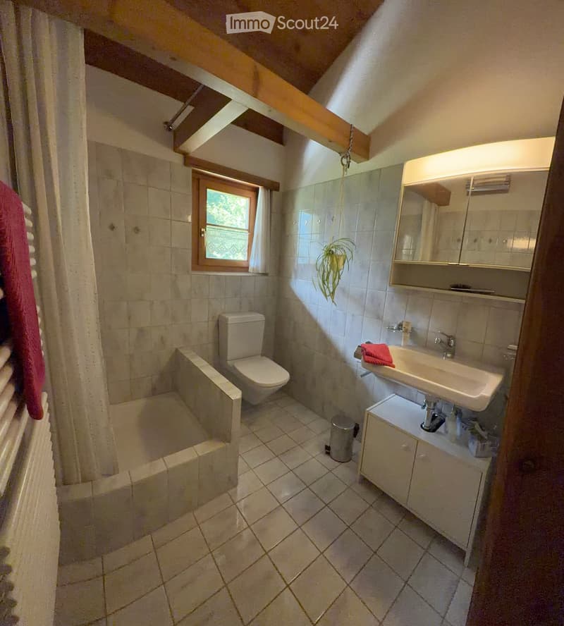 Cozy bathroom with tiled walls and floor, featuring a toilet, sink with mirror, bathtub, and a small window with a plant on the windowsill.