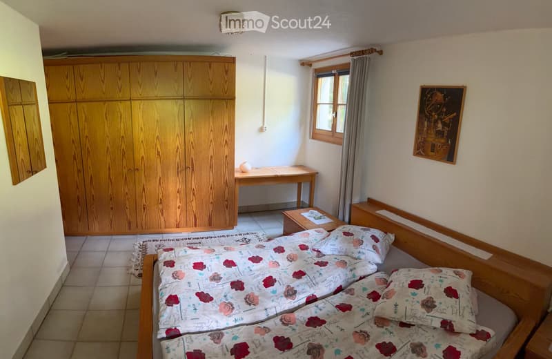 Cozy bedroom with a double bed, wooden wardrobe, nightstand, small table, and a framed picture on the wall.