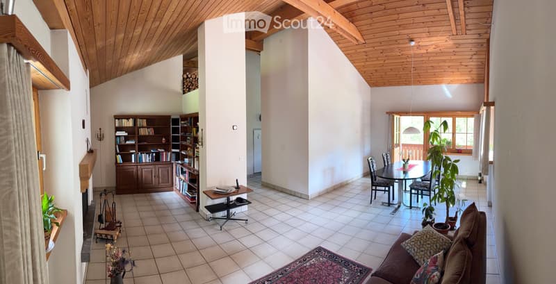 Spacious living and dining area with wooden ceiling beams, tiled floor, and large windows. Features a sofa, dining table with chairs, bookshelf, and decorative plants.