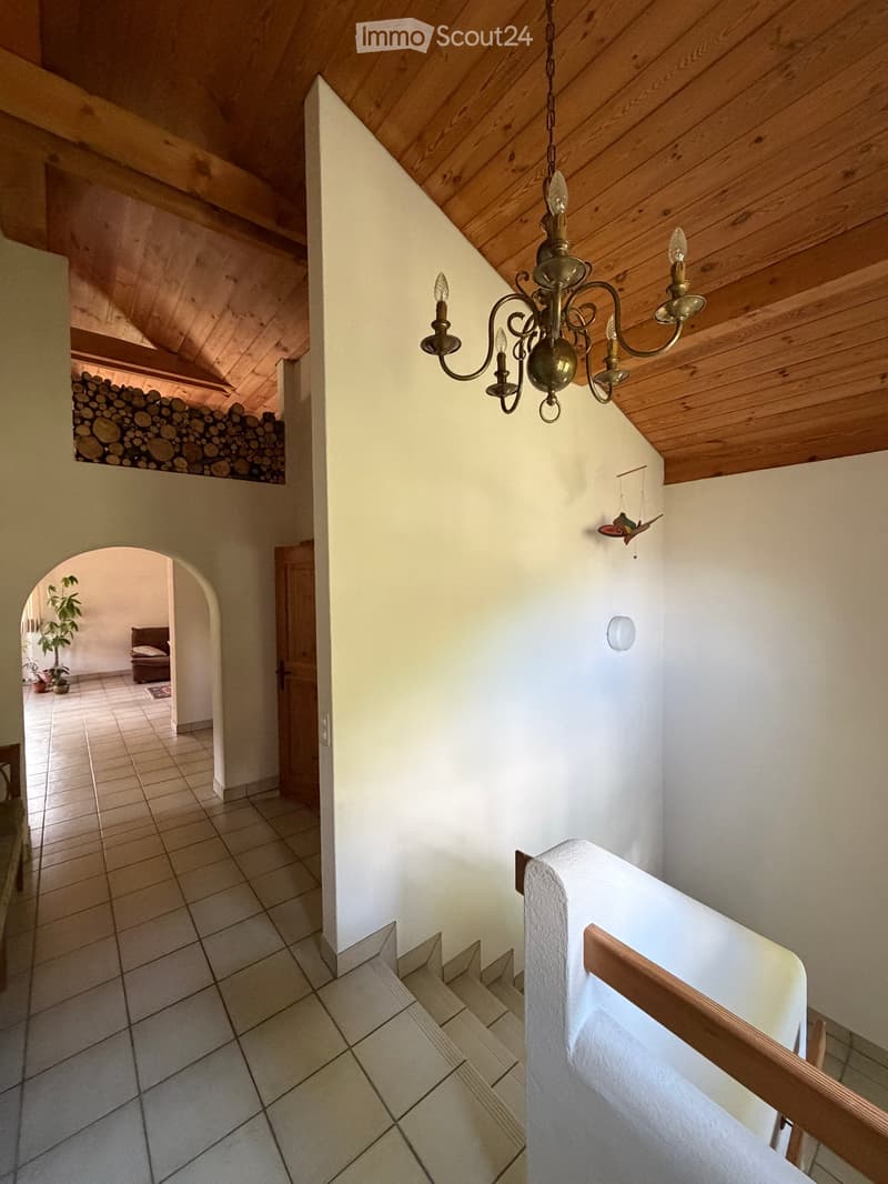 Staircase with wooden railing, chandelier, and visible storage of firewood in the background. The hallway has tiled flooring and white walls.