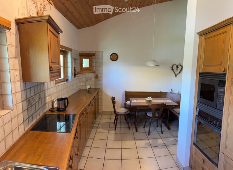 Bright kitchen with wooden cabinets, tiled backsplash, and a dining area with a table and chairs. Features include a microwave, oven, electric kettle, and a clock on the wall.