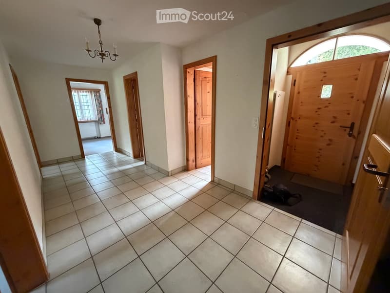 Hallway with tiled floor, wooden doors, and a chandelier hanging from the ceiling. Visible rooms include a windowed area with curtains and a partially visible room with a dark rug and shoes.