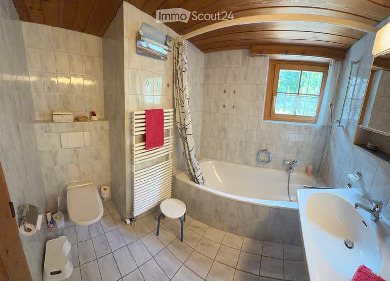 Bathroom with a toilet, bathtub, sink, and a towel rack. The room features tiled walls and a window with a curtain. A small stool is placed near the toilet, and a mirror is mounted above the sink.