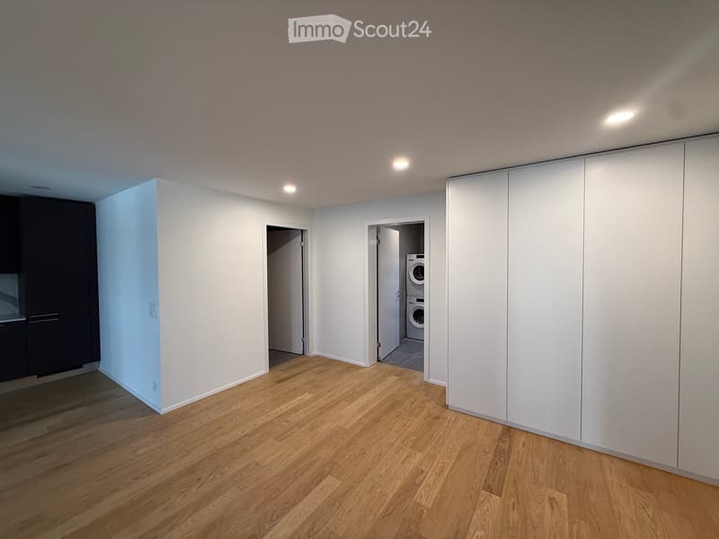 Empty room with light wooden flooring, white walls, and recessed lighting. A closet with white sliding doors is visible, and an open doorway leads to a small area with a washing machine.