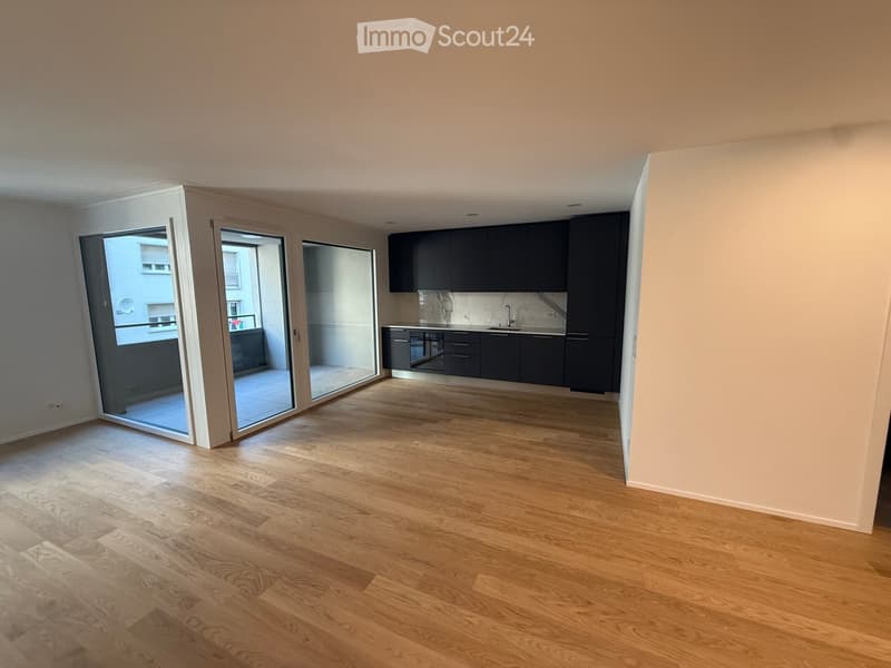 Empty room with wooden flooring, large sliding glass doors leading to a balcony, and a small kitchen area with dark cabinets and a sink.