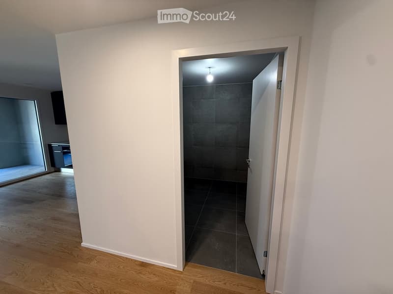Empty hallway with white walls, wooden flooring, and a partially open door leading to a dark tiled room.