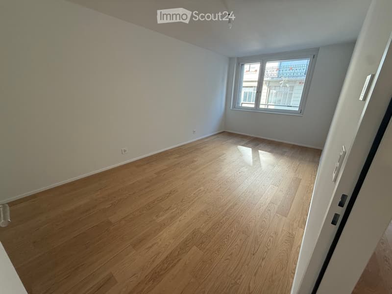 Empty room with wooden flooring and white walls, featuring a large window with a view of a balcony and neighboring building.