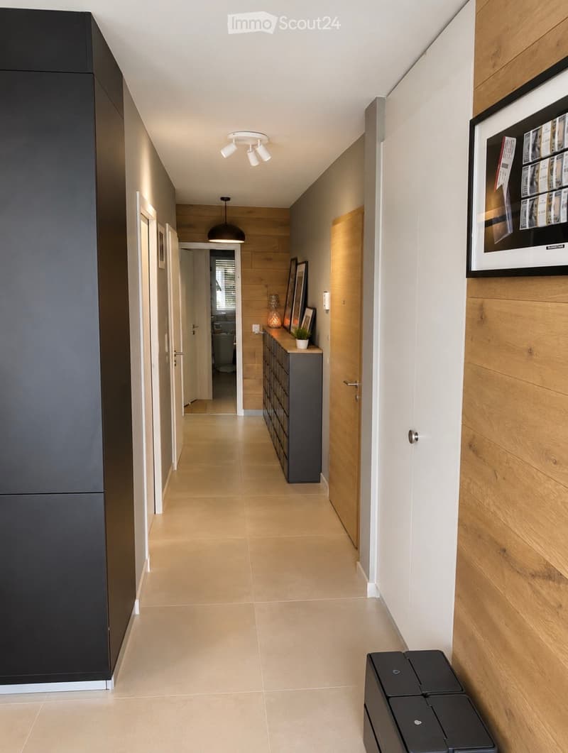 Modern hallway with light-colored tiles, wooden paneling on one wall, and built-in storage unit with framed artwork above it. Features recessed ceiling lights and a small plant on the storage unit.