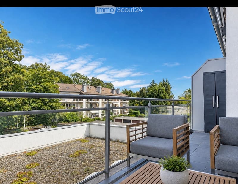 Balcony with modern grey sofa, wooden table, and potted plant, overlooking a green residential area with trees and buildings.