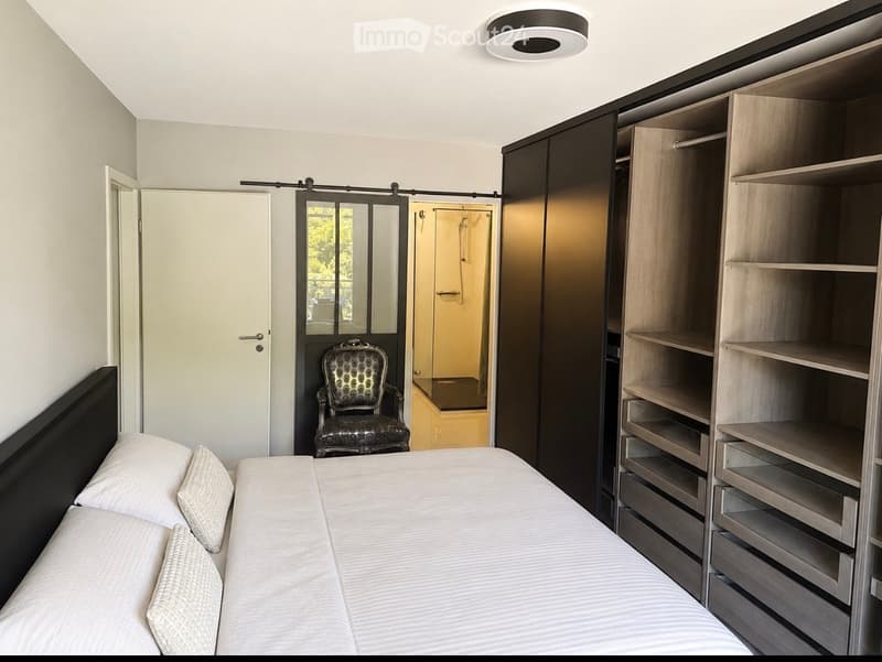 Modern bedroom with a large bed, patterned chair, built-in wardrobe with shelves and drawers, and a sliding door leading to a shower area.
