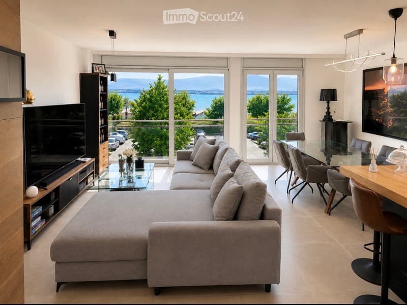 Modern living room with large windows offering a scenic view, featuring a grey sectional sofa, glass coffee table, dining area with chairs and table, and a TV stand with a screen.
