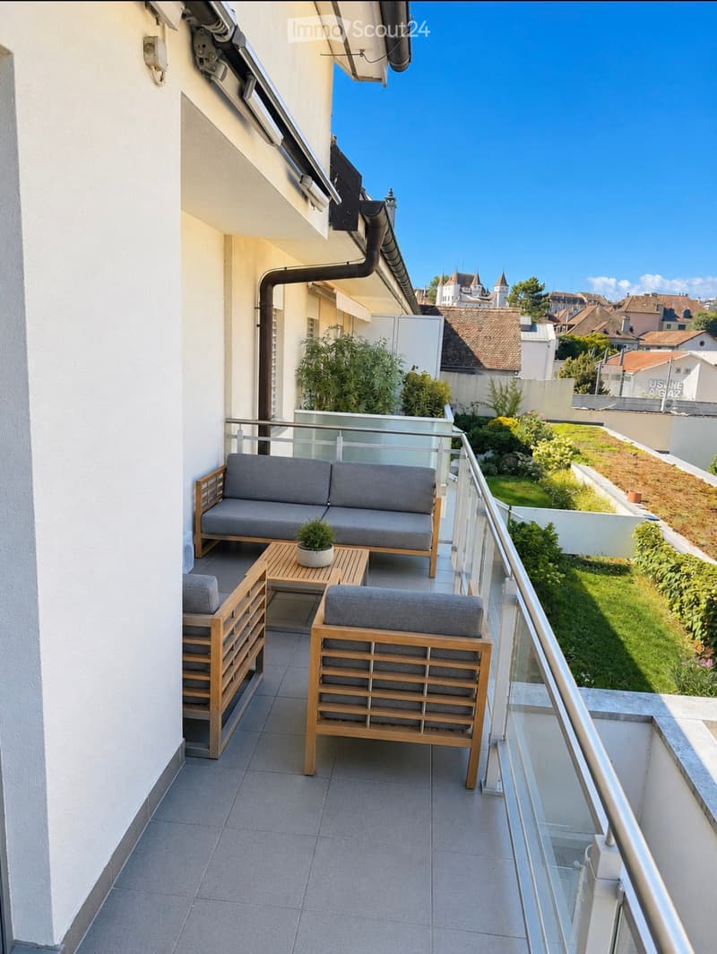 Balcony with wooden furniture including a sofa, table, chairs, and a small potted plant, overlooking a landscaped garden area.