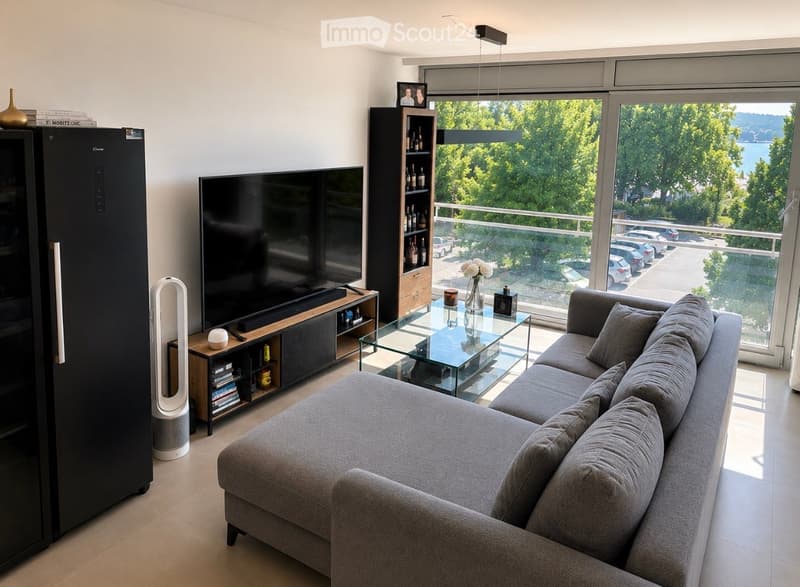 Modern living room with large windows offering a scenic view, featuring a grey sectional sofa, glass coffee table, flat-screen TV, and a sleek black refrigerator.