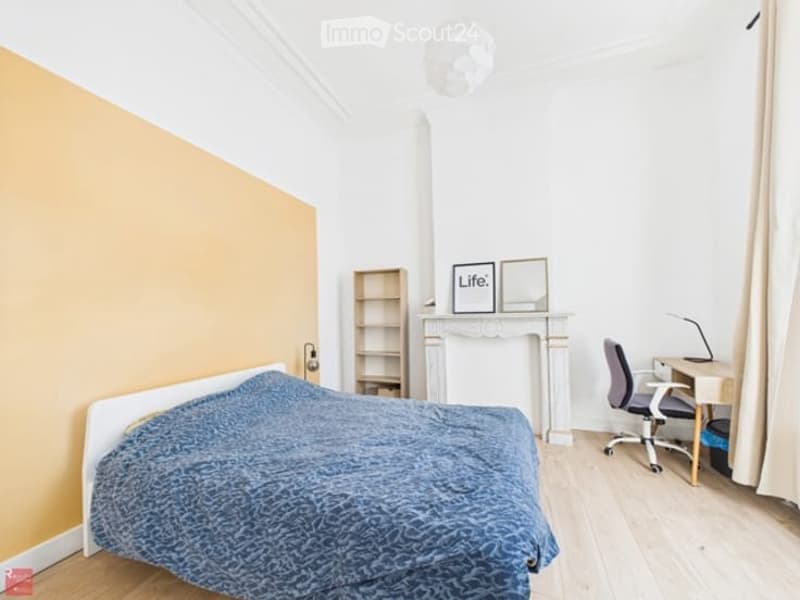 Cozy bedroom with a blue bedspread, a fireplace with a mirror above it, a wooden shelf, a desk with a chair, and a lamp on the desk.