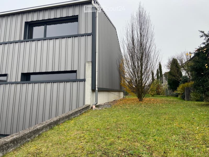 Modern, single-story building with grey metal siding, large windows, and a raised ground floor. The surrounding area includes a grassy lawn and trees.