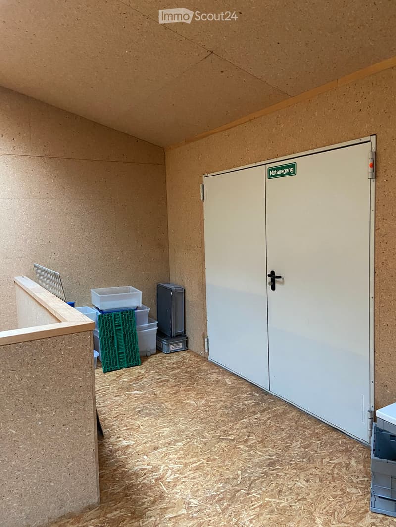 A small, unfurnished room with wooden walls and a wooden floor, featuring a door labeled 'Notausgang' and a few storage items including a green crate, a white plastic bin, and a small refrigerator.