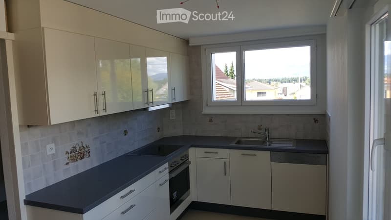 Modern kitchen with white cabinets, dark countertops, and stainless steel appliances including a sink, oven, and dishwasher. Large window offers a view of the surrounding area.