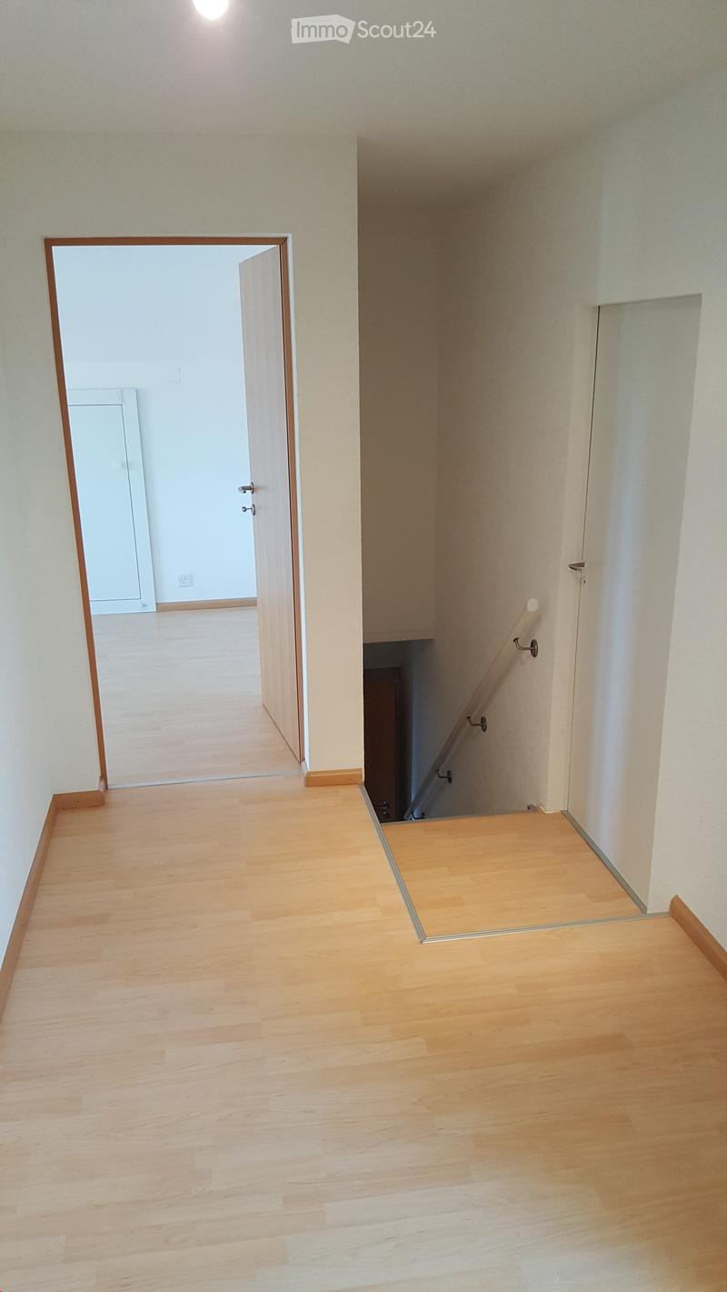 Empty hallway with light wooden flooring, white walls, and a staircase leading upwards. An open doorway leads to another room.