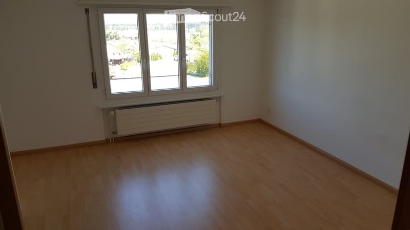 Empty room with wooden flooring, white walls, and a large window with a view of the outside. A radiator is visible below the window.