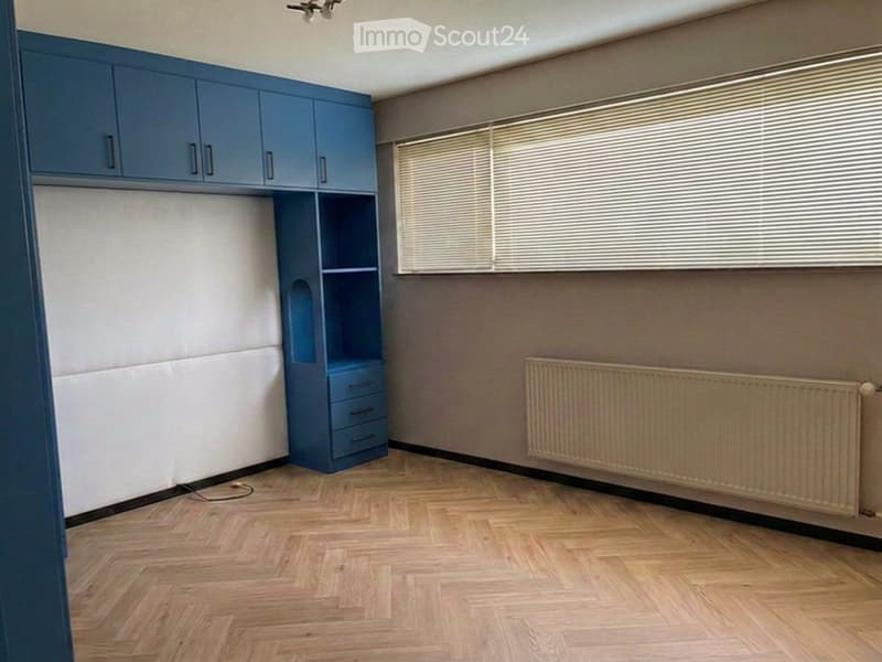 Empty room with wooden flooring, blue built-in storage unit, radiator, and window with blinds.