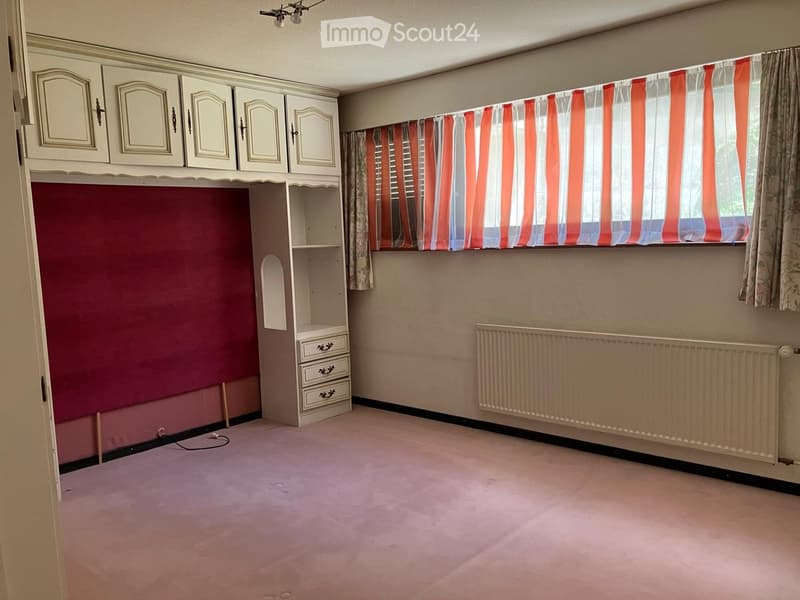 Empty room with pink carpet, white built-in cabinets, a radiator, and striped curtains on the window.