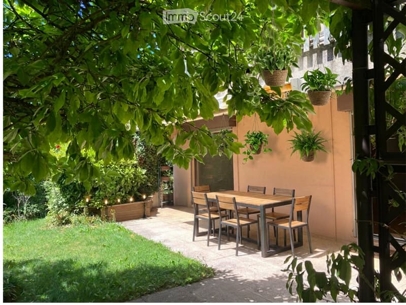 A shaded outdoor dining area with a wooden table and chairs, surrounded by greenery and hanging plants.