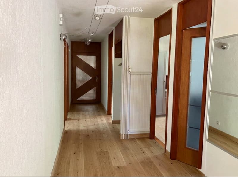 A long, narrow hallway with wooden flooring, white walls, and several wooden doors with glass panels. The hallway features a radiator on one side and a ceiling light fixture.