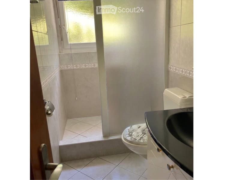 Bathroom with a toilet, sink, and shower area. The shower has a glass door and a window above it. The walls are tiled with a decorative border near the floor.