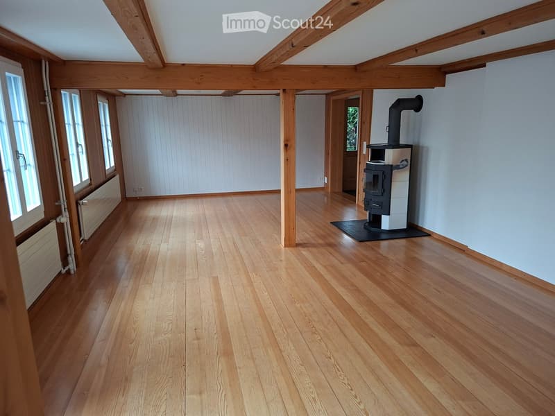 Empty room with wooden flooring, exposed wooden beams, white paneled walls, and a wood-burning stove with a chimney.